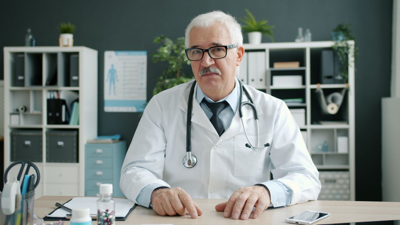 Offerings Senior doctor in an office setting, with medical equipment and files in the background.